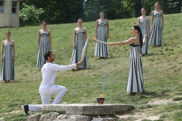 扮演最高女祭司的演員瑪麗·米娜（前右）點(diǎn)燃第一棒火炬手——希臘運(yùn)動(dòng)員、東京奧運(yùn)會(huì)男子賽艇項(xiàng)目金牌得主斯特凡諾斯·恩圖斯科斯手中的火炬。新華社記者 趙丁喆 攝