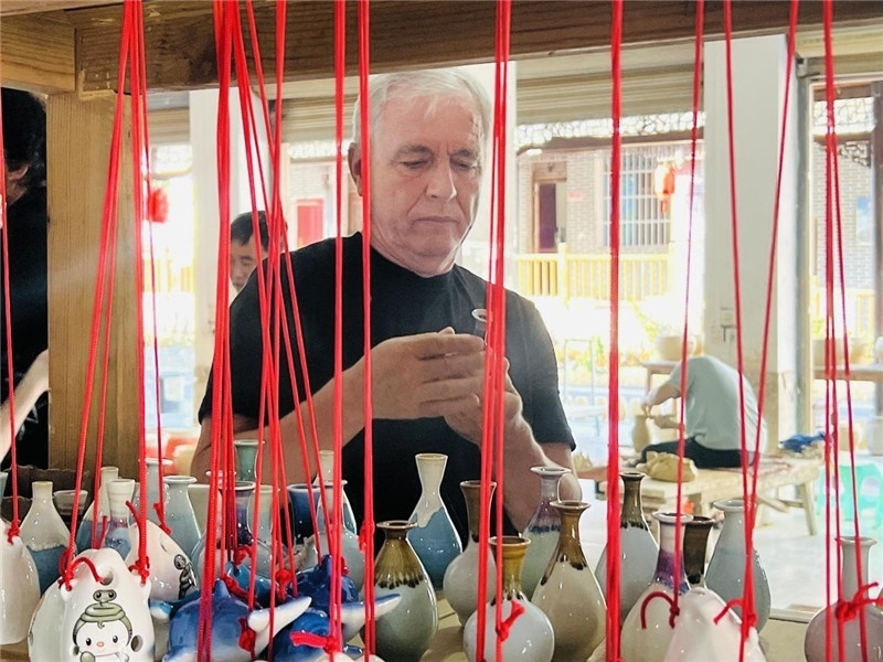鄉(xiāng)愁如同一根根紅線(xiàn)，將游子與故鄉(xiāng)永遠(yuǎn)地牽連到一起?；逄账囄幕瘎?chuàng)意一條街的母氏陶藝館內(nèi)，紅線(xiàn)吊著陶瓶，思緒游走萬(wàn)里。摩爾多瓦“信息標(biāo)簽”通訊社社長(zhǎng)亞歷山大·塔納斯端詳著館內(nèi)展示的陶藝作品，并挑選出心儀的小擺件帶回摩爾多瓦，也帶走花茂村的“鄉(xiāng)愁”。（張亞欣攝）
